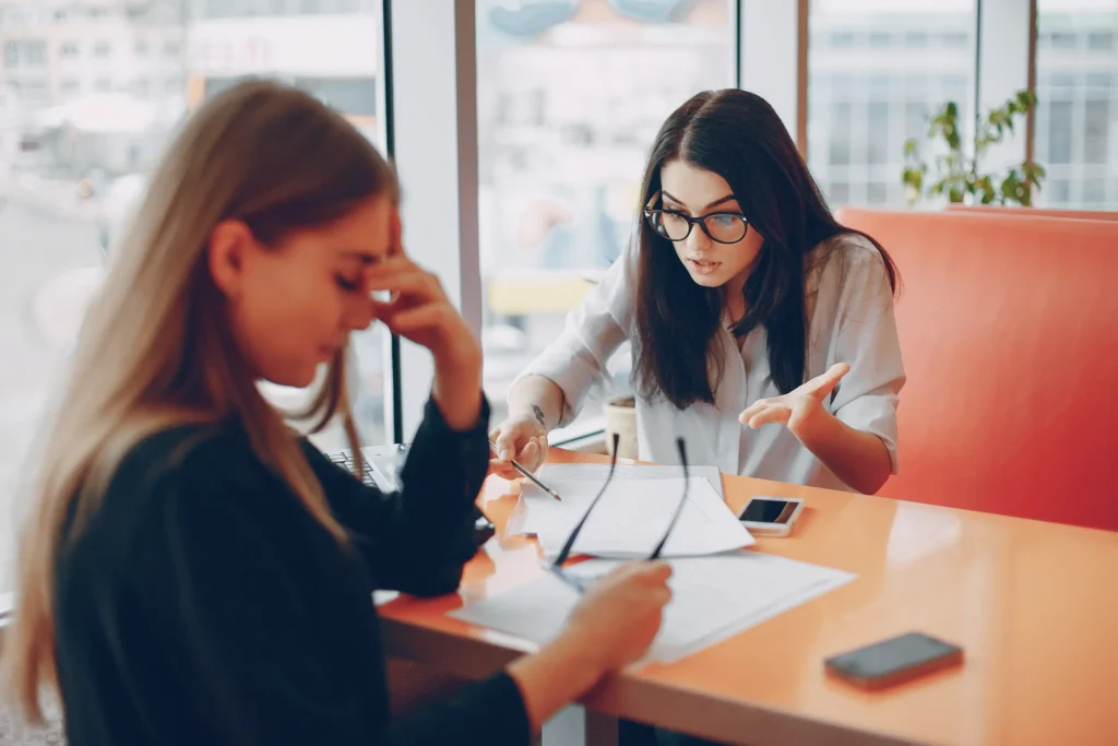 Dos mujeres durantes una entrevista labora, una muestra nerviosismo sujetándose la cabeza, mientras la otra revisa su curriculum y le habla expresivamente brindando orientación.