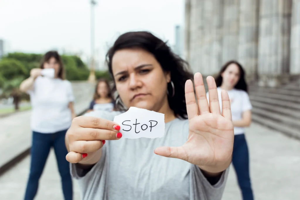 Mujer mostrando la palma de la mano en señal de alto y sosteniendo un cartel que dice “STOP”, con otras mujeres desenfocadas al fondo, simbolizando la detección de la discriminación laboral.