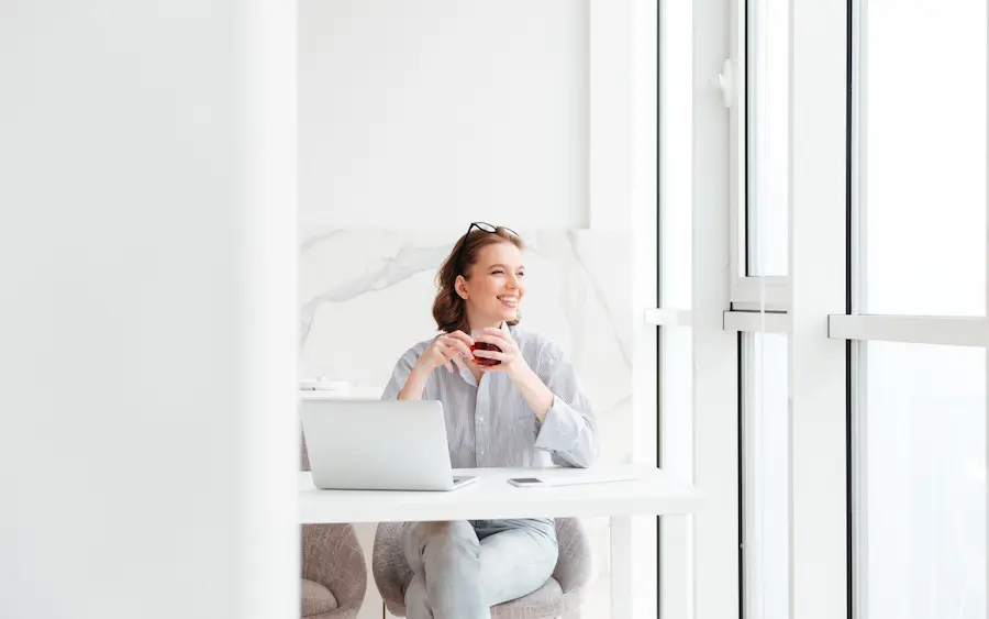 Mujer sonriendo con una taza de café frente a su laptop, mirando por la ventana con actitud reflexiva y tranquila.