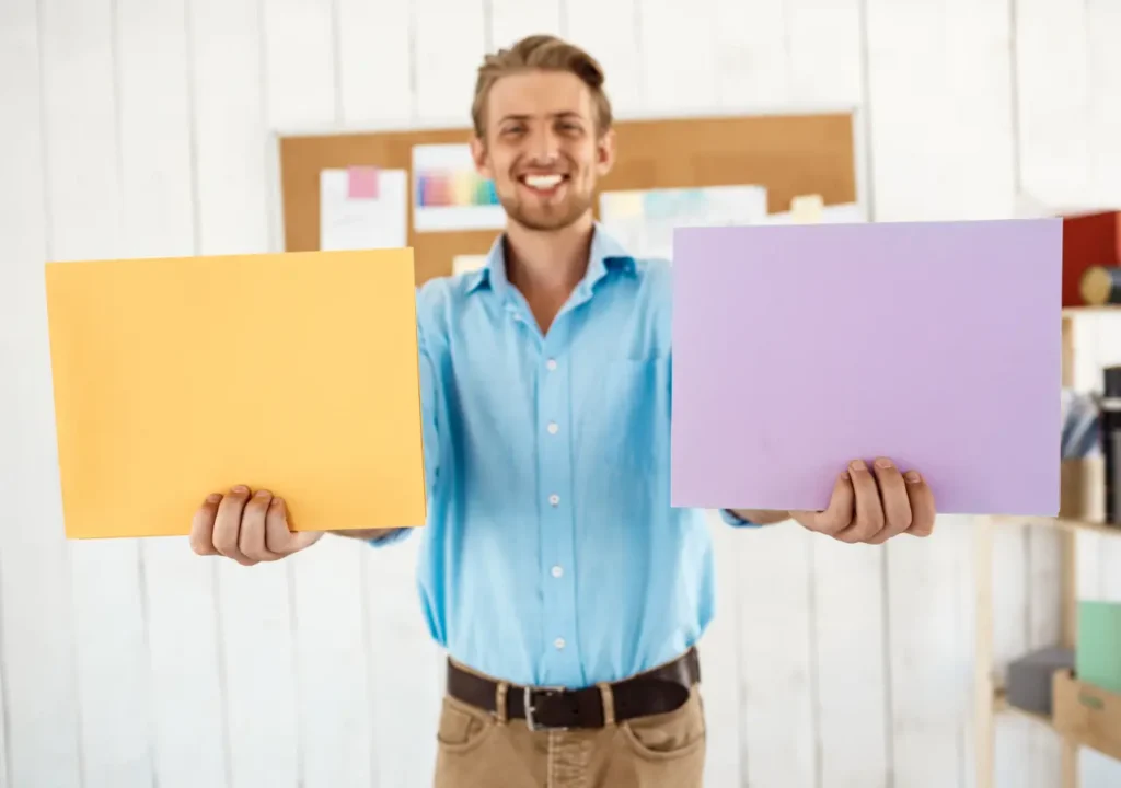 Hombre sonriente sosteniendo dos carpetas de colores diferentes, simbolizando la elección entre opciones laborales y la búsqueda de la empresa adecuada para su perfil profesional.